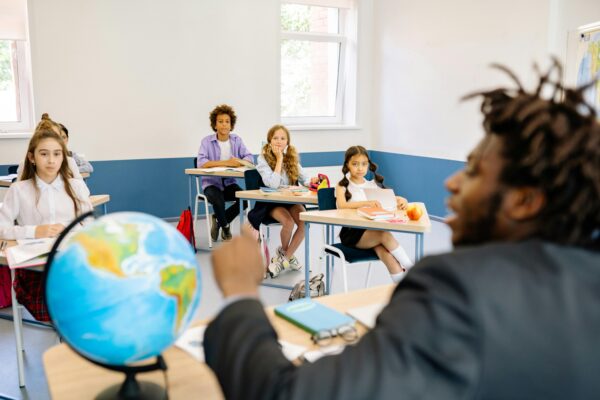 Children in a classroom with a teacher pointing at a globe, representing education and child Health in a positive school environment.
